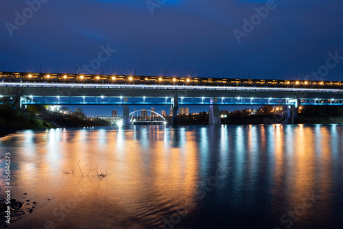THE HIGH LEVEL BRIDGE AT 100, in a beautiful sunrise with shades of blue, yellow and red lights, over a river of blue water with reflections and swept clouds, stones and yellow leaves in edmonton city