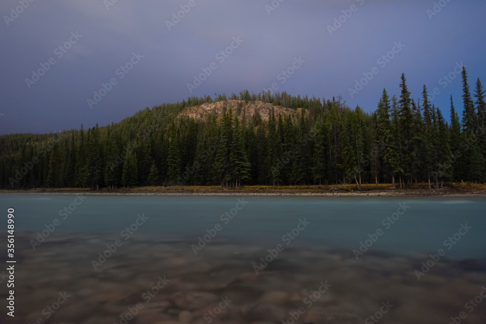 Night photography of Maligna Lake in Jasper National Park Alberta Canada. with a blue night sky, and a turquoise blue lake and rocks on the shore, with a green background of pine trees.
