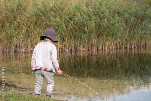 Child playing in nature. Rear view of one boy standing beside the lake pretending to fish with stick in hand in Switzerland.