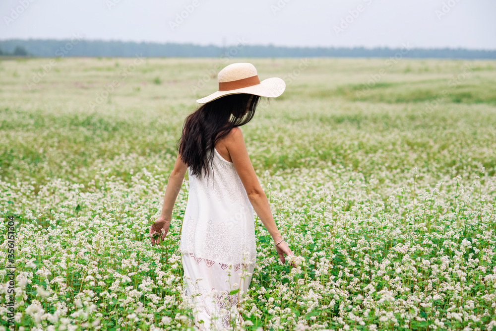 A girl with dark hair in a white dress and a straw hat walks on a white flower field. Summer rural landscape. Tender brunette portrait