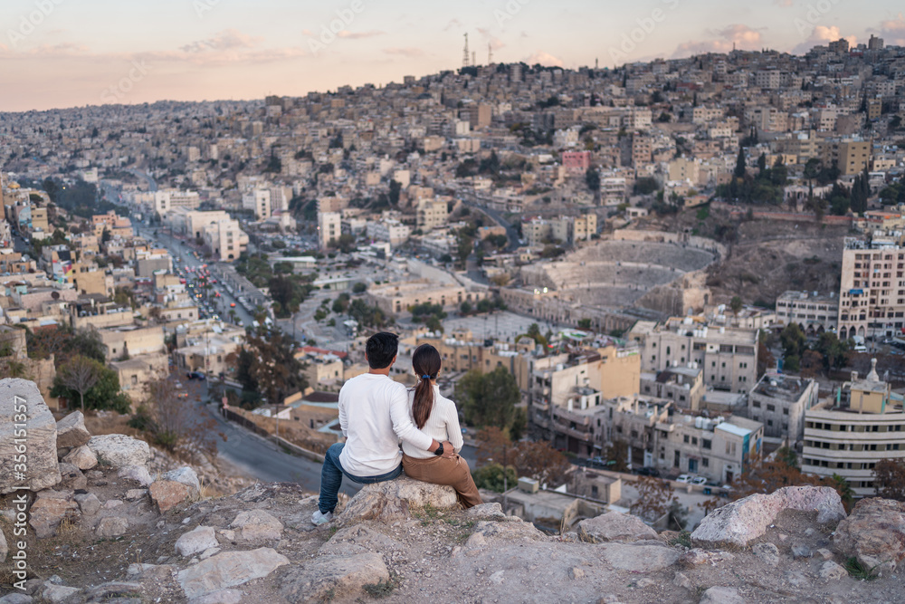 Young couple in love sitting on rock and enjoying top view of Amman city at sunset, Jordan