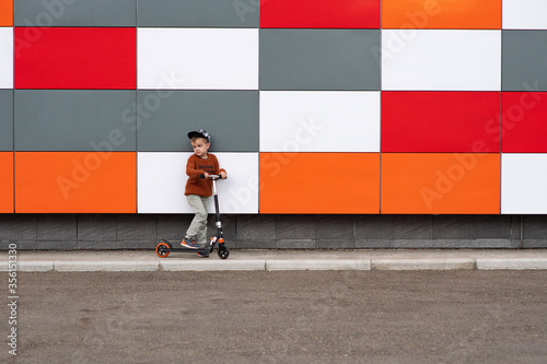 A boy in an orange sweater and light jeans rides a scooter. Children's vehicle. Stylish children's clothing in bright colors. Active lifestyle of modern children. European appearance, 6 years old © Ekaterina