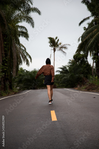 Girl in a brown leather jacket is on a paved road with a yellow stripe amongst the palm trees © vgatto