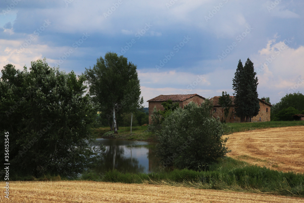 Come in un dipinto dell’ottocento: panorama collinare con case rurali, laghetto e campo di grano appena mietuto sotto un cielo minaccioso e temporalesco