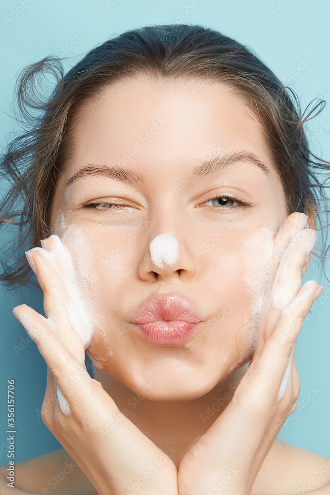 Face masks are the perfect treatment to hydrate skin, remove excess oils and improve the appearance of pores. A young brown-haired lady is applying white bubbling mass on her face and sending a kiss.