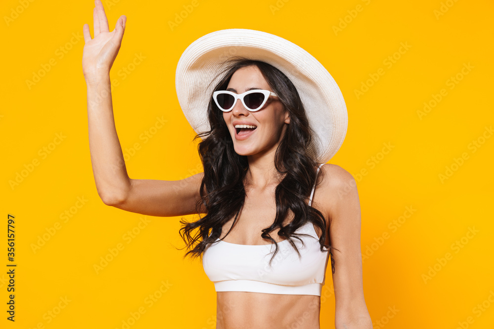 Image of stylish woman in swimsuit and hat smiling and waving hand