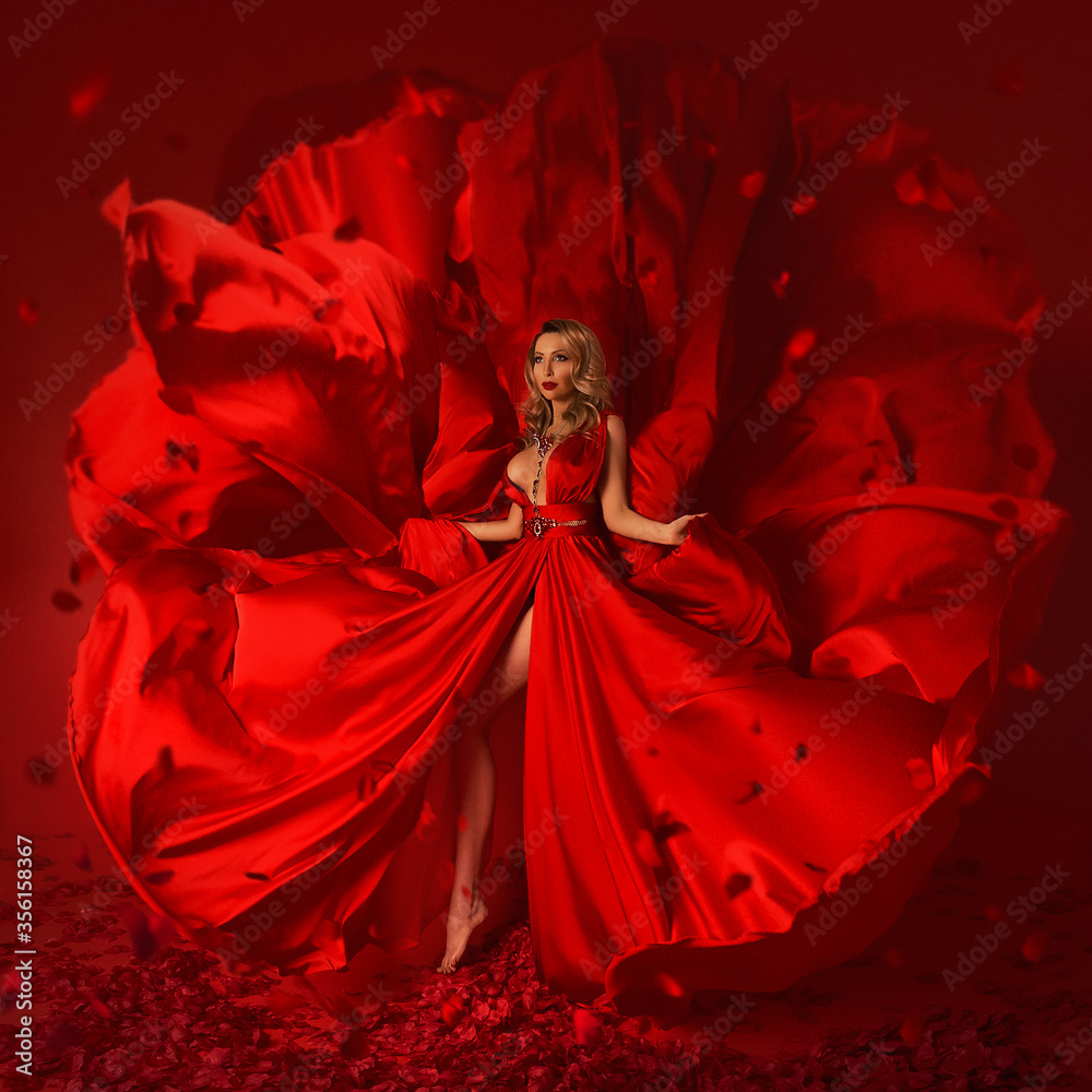 A gorgeous blonde girl in a luxurious flying red silk dress with a deep slit standing barefoot on a red background with rose petals. A gorgeous blonde girl in a luxurious flying red silk dress with a deep slit standing barefoot on a red background with rose petals.