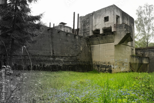 A semi black and white photo of the exterior of an old dilapidated dam built by the Nazis during World War Two with a lush meadow full of flowers and herbs in the foreground seen on a spring day