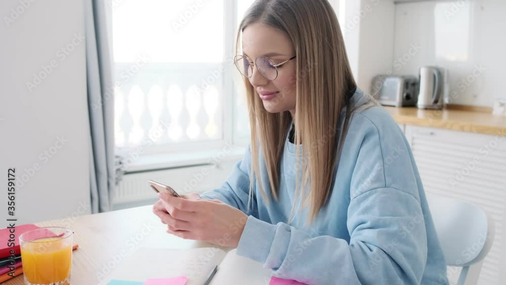 A focused young blonde woman wearing glasses is typing on her smartphone sitting in the kitchen at home A focused young blonde woman wearing glasses is typing on her smartphone sitting in the kitchen at home