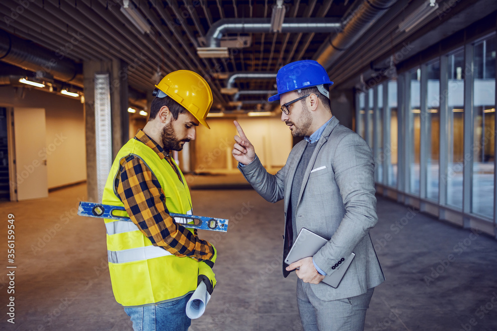 Angry caucasian businessman in suit and helmet on head arguing with irresponsible construction worker. Building in construction process interior. Angry caucasian businessman in suit and helmet on head arguing with irresponsible construction worker. Building in construction process interior.