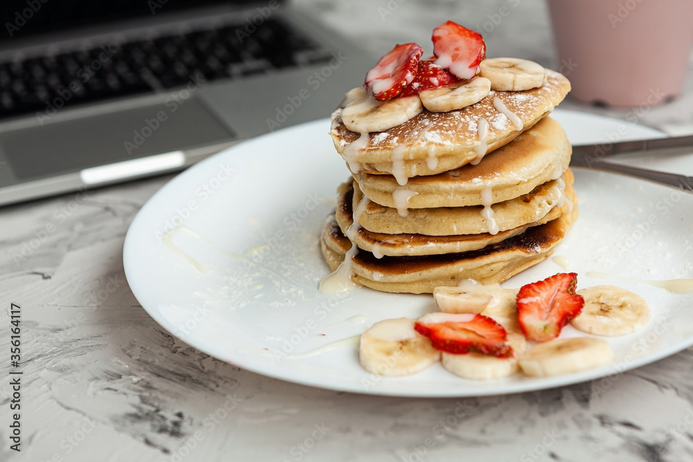 A stack of pancakes with fresh strawberries on a white plate on a light background. There is a laptop next to it. Work at home. Horizontal orientation. Homemade american pancakes. Copy space. A stack of pancakes with fresh strawberries on a white plate on a light background. There is a laptop next to it. Work at home. Horizontal orientation. Homemade american pancakes. Copy space.