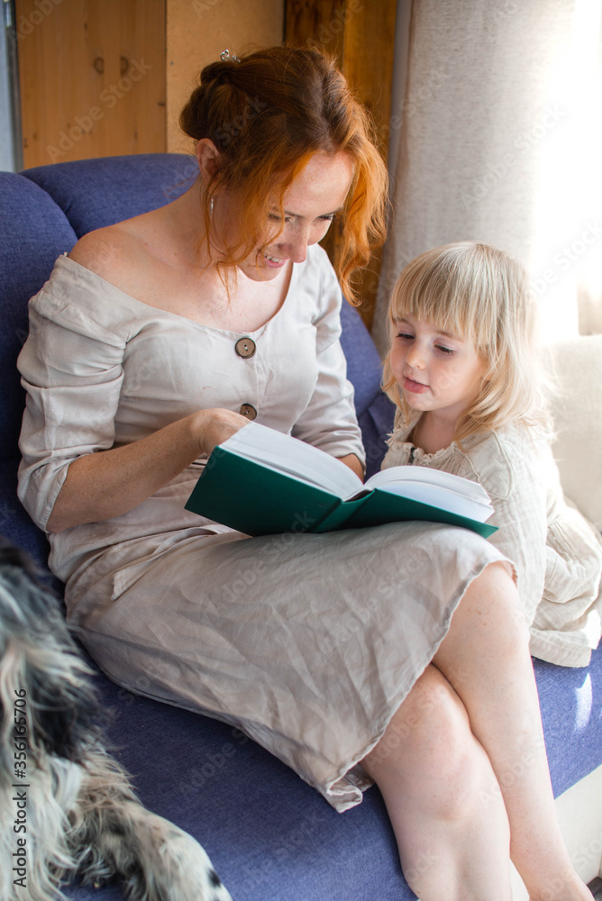 A beautiful young woman and her little cute daughter are sitting on a blue sofa and reading their favorite book with their dog at home.