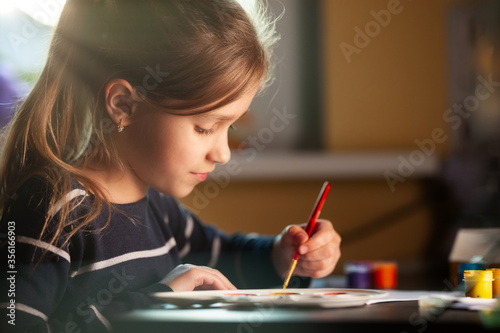 Side view of charming little caucasian smiling schoolgirl writes while sitting at table. Little cute girl draws picture. Concept of teaching children during self-isolation