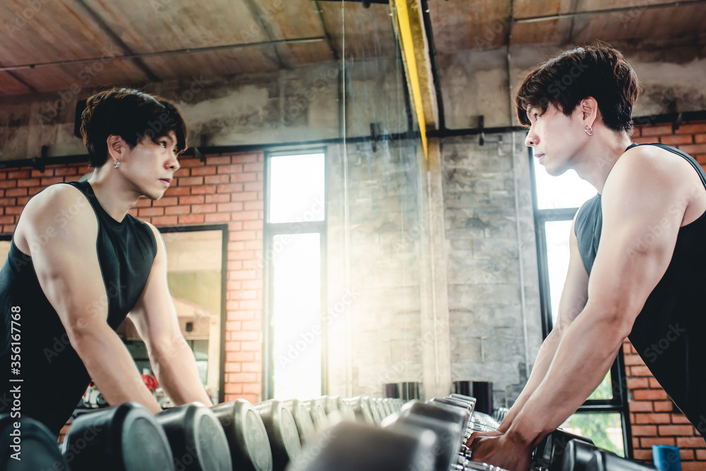 A young Asian man raised a dumbbell to exercise with a dumbbell in the gym. To strengthen the arm muscles Look in the mirror at the fitness center. Concept: Fitness man with dumbbell in gym. A young Asian man raised a dumbbell to exercise with a dumbbell in the gym. To strengthen the arm muscles Look in the mirror at the fitness center. Concept: Fitness man with dumbbell in gym.