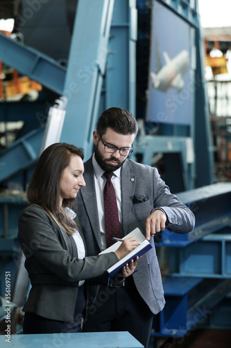 Portrait of a solid businessman with his secretary holding blue notebook, talking about factory financial report in a airplane manufactory. Factory managers controlling work process in a factory