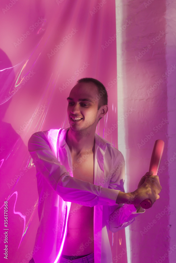Portrait of the Young Handsome emotional man in an unbuttoned white shirt with bat breaks the ball. Neon Room. Old 90's retro style. Reflect of neon words near male. Selective focus. Portrait of the Young Handsome emotional man in an unbuttoned white shirt with bat breaks the ball. Neon Room. Old 90's retro style. Reflect of neon words near male. Selective focus.