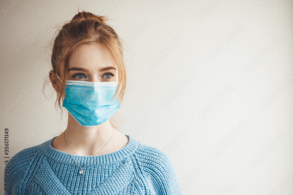 Ginger girl with freckles wearing medical mask and knitted blue sweater is posing on a white studio wall with free space Ginger girl with freckles wearing medical mask and knitted blue sweater is posing on a white studio wall with free space