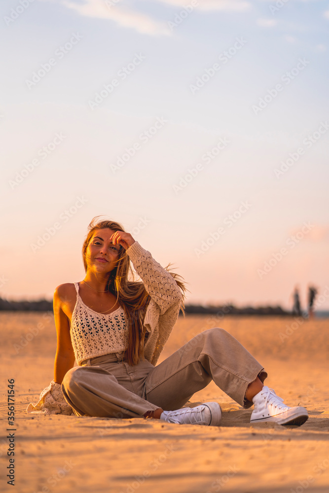 Summer lifestyle, a young blonde with straight hair, in a small wool sweater and corduroy pants sitting on the beach. Enjoying a summer afternoon at sunset sitting on the sand