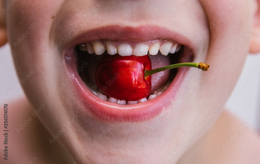Obraz premium The child holds a delicious cherry in his mouth. Beautiful red berry. The child smiles.