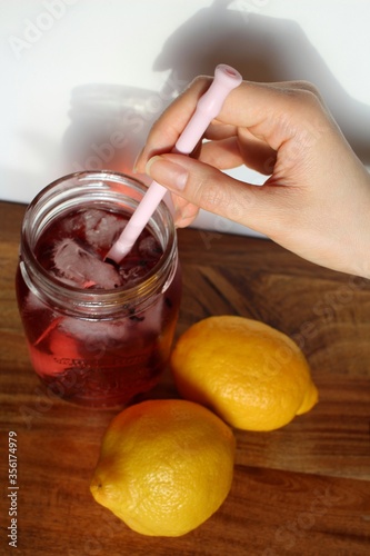 Woman's hand holding a pink reusable straw in a glass mason jar full of strawberry lemonade on a textured wooden table with ripe yellow lemons close by. The perfect tropical vacation drink.