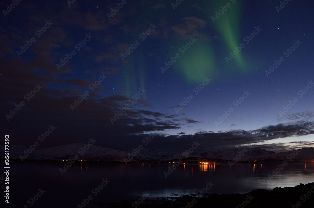 faint aurora borealis over calm fjord at night with lights in the background