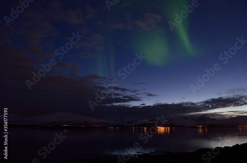 faint aurora borealis over calm fjord at night with lights in the background