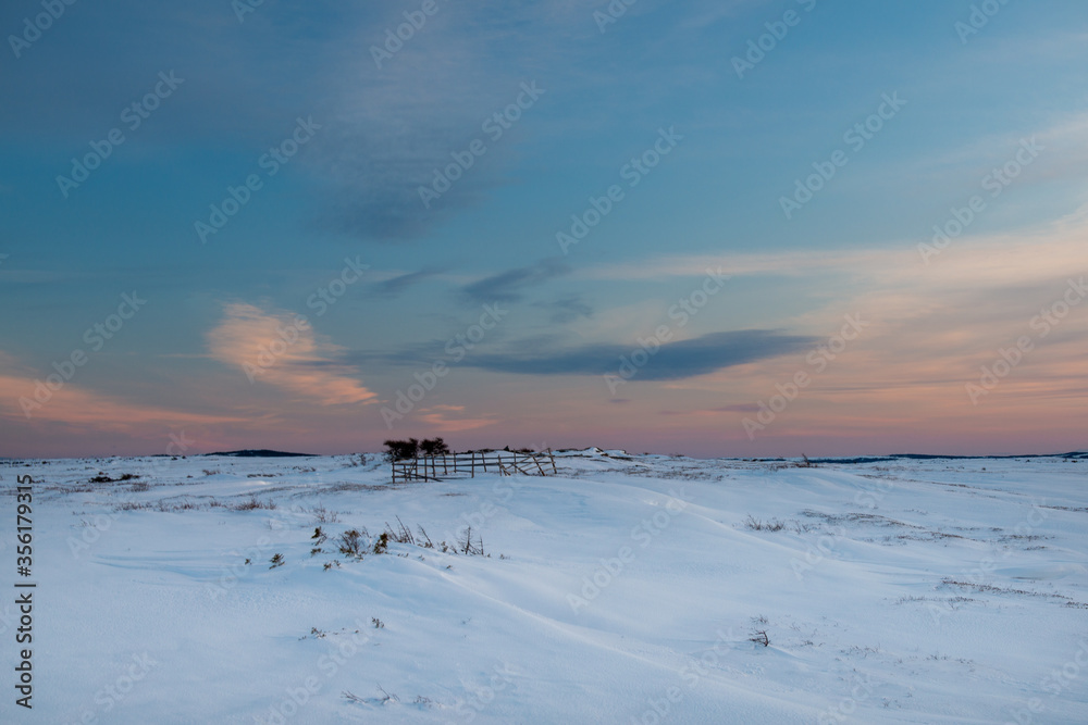 Barren land with white snow covering the marsh, There's a wooden square fence or pen with a small abandoned farm building. There's a pink setting sky in the background with low lying clouds.