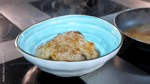 Chef takes hot fried pork meat, chop from the pan with cooking tongs and put it in blue bowl with potatoes inside. Delicious meal cooking procees. Induction cooker. photo