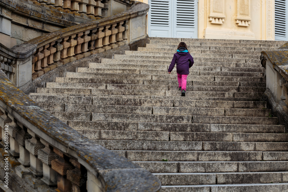 Unrecognisable little girl, modern-day lonely princess walking up majestic curved stairs of empty castle in Europe. Child alone visits historic European site walks up large, decorated flight of stairs