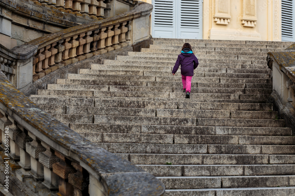 Unrecognisable little girl, modern-day lonely princess walking up majestic curved stairs of empty castle in Europe. Child alone visits historic European site walks up large, decorated flight of stairs Unrecognisable little girl, modern-day lonely princess walking up majestic curved stairs of empty castle in Europe. Child alone visits historic European site walks up large, decorated flight of stairs
