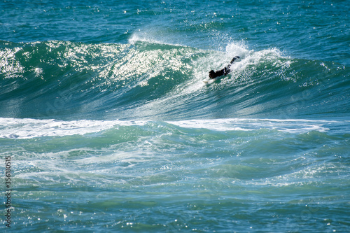 A male swims among large blue ocean waves while trying to stand on a surfboard. The waves have a spray coming off the curls. The cold Atlantic Ocean has a blue to green tint to the water. 