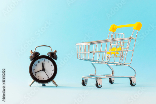 Alarm clock and miniature shopping cart on light blue background. Purchasing time. Buying time. Business concept.