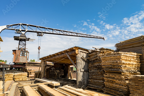 Sawmill with boards and sawn trees near a large old crane with rust under a blue sky with white clouds in summer. Deforestation and logging.