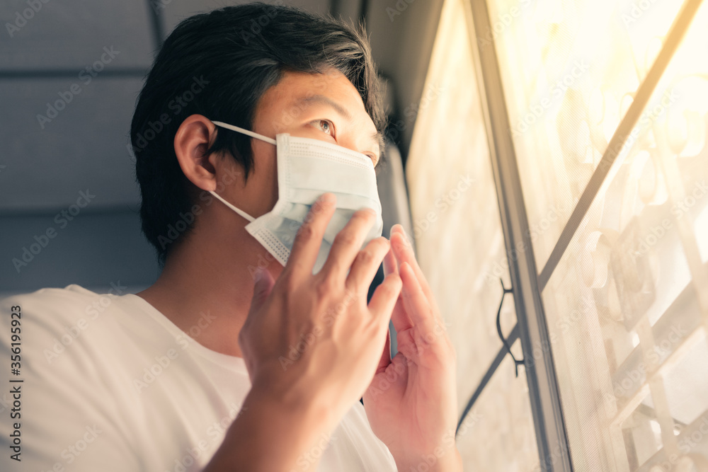 The young man was detained at home during the coronavirus outbreak, an Asian man standing by the window and wearing a mask to prevent germs and to keep away from society