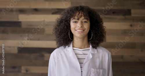 Konstfotografi CU Young Hispanic woman in white lab coat looking to camera, expressing happiness and confidence