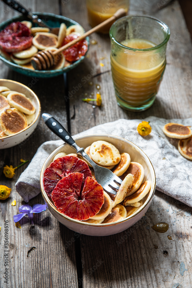 Ceramic bowl with mini pancakes, blood orange slices and honey on old wooden table.