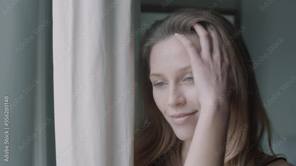 Woman going through her hair with hand while standing next to a window Woman going through her hair with hand while standing next to a window