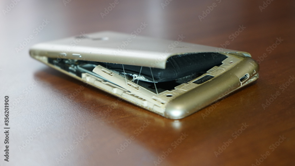 Swollen smartphone battery. Damaged smartphone with a faulty battery. Damaged phone on a wooden background. Swollen smartphone battery. Damaged smartphone with a faulty battery. Damaged phone on a wooden background.