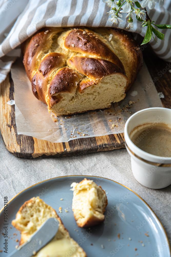 Homemade braided bread (challah) half covered with a towel, slice of it on plate, butter, cup of coffee and bouquet on table. Homemade braided bread (challah) half covered with a towel, slice of it on plate, butter, cup of coffee and bouquet on table.