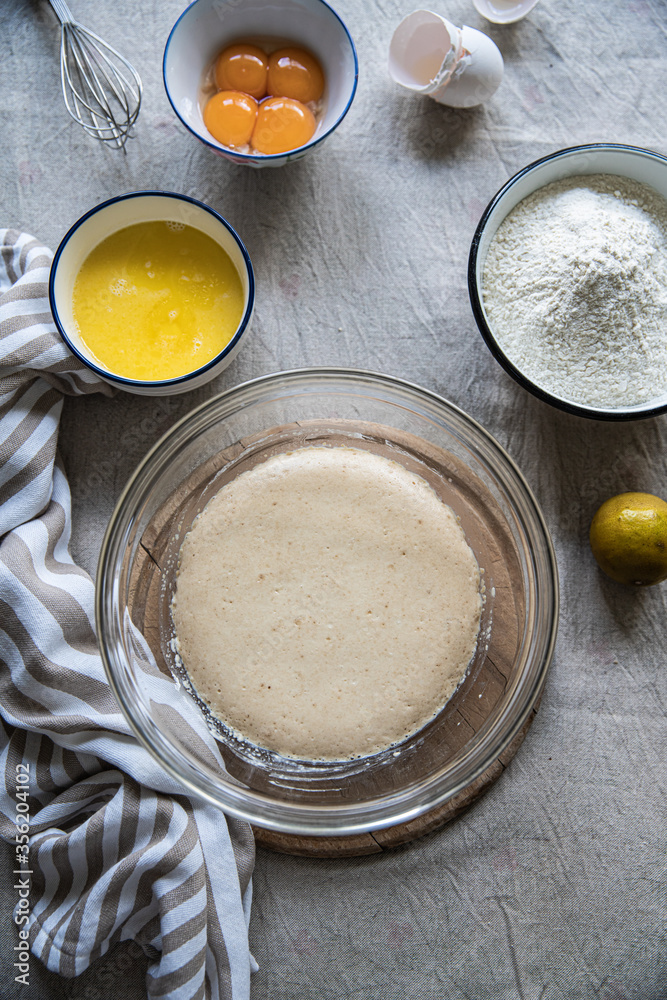 Step by step process of making yeast bread. Top view of ingredients: bowls with yeast starter, melted butter, egg yolks and flour, lemon, egg shells and towel on linen tablecloth. Step by step process of making yeast bread. Top view of ingredients: bowls with yeast starter, melted butter, egg yolks and flour, lemon, egg shells and towel on linen tablecloth.
