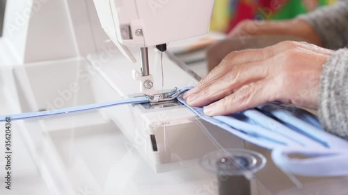 Elderly woman stitching a strap on a blue cotton face mask with a sewing machine. 