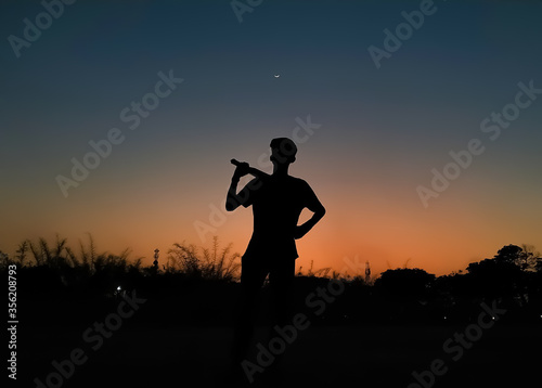 Silhouette of a boy playing cricket in the evening with the crescent moon in the colorful sky after sunset as wallpaper background. Gully cricket. Cricket wallpaper. India. Cricket during pandemic.