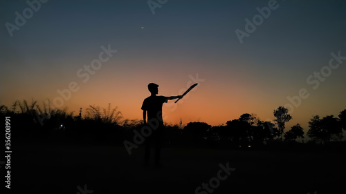 Silhouette of a boy playing cricket in the evening with the crescent moon in the beautiful sky full of variety of colors after sunset as wallpaper background. Gully cricket is very popular in India.
