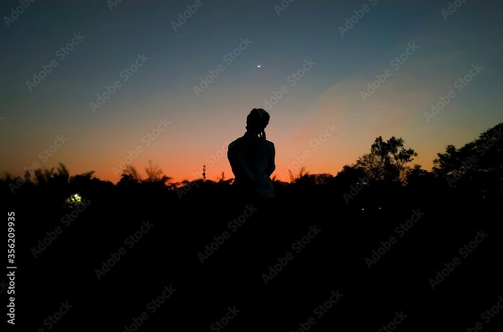 Silhouette of a man in deep thought and questions with hand on his cheeks in the evening with the crescent moon in the colorful sky after sunset as background. Inspirational. Declutter mind concept.