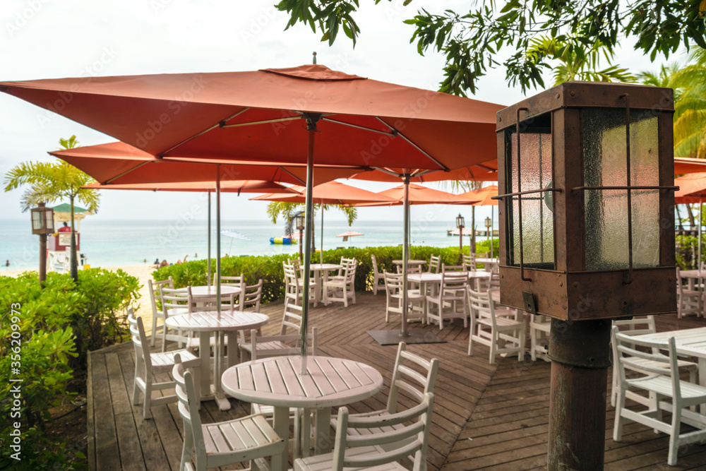 Montego Bay, Jamaica. Restaurant terrace on the beach with large orange patio umbrellas and empty tables and chairs. Ocean in background.