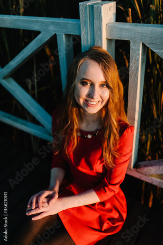 Portrait of a beautiful girl sitting in autumn in a red dress against the background near the fence on the nature. upper half. look straight ahead. Close up. headshot.