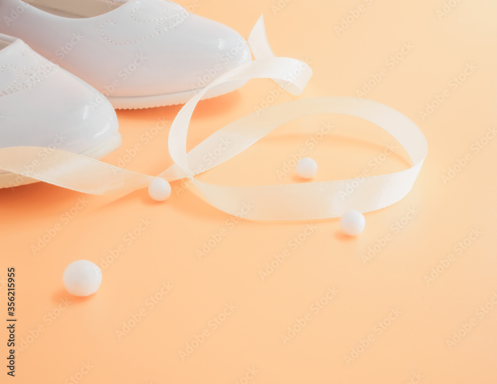 Pair of white baby lacquered girls shoes with beige ribbon and white balls at peach background with place for text Pair of white baby lacquered girls shoes with beige ribbon and white balls at peach background with place for text