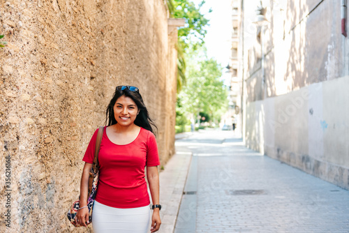 Smiling latin girl walking next to wall looking at camera on sunny day.