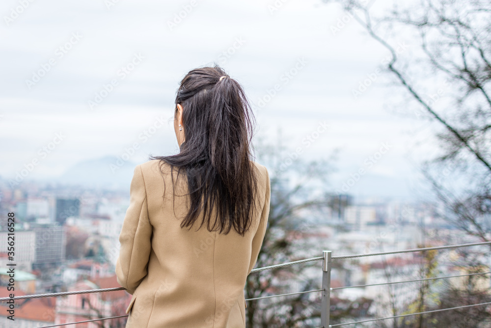 Young business woman with long dark hair looking down on Ljubljana city wearing beige coat. Winter season on Ljubljana Castle, Slovenia. Shallow depth of field