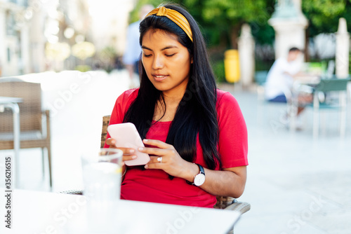 Young brunette Latin woman with long hair sitting on bar terrace looking at a mobile phone